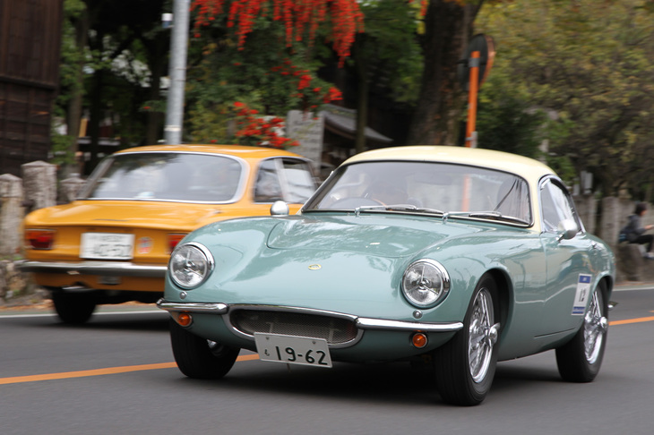 正門を出てラリーコースを行く1962年「ロータス・エリートSr2」と、イベントを訪れたギャラリーのクルマであろう「アルファ・ロメオ1300GTジュニア」が、偶然すれ違った。