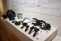 「ルノーR25」のエグゾーストパイプ、ブレーキディスク、ブレーキペダル、タイヤやホイールなどのパーツ類。壁面に掛けられたパネルには各パーツの解説が記されている。