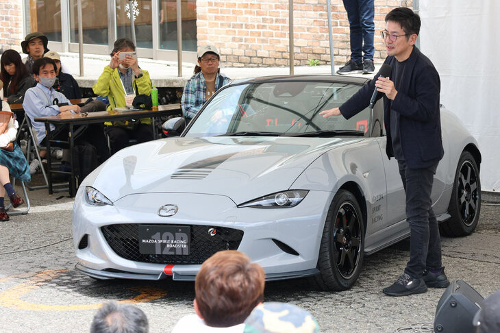 「マツダ スピリット レーシング・ロードスター12R」の特徴を解説する諌山慎一さん。仕様は1種類で、ボディーカラーは「エアログレイ」のみ。その狙いはスーパー耐久参戦車両との共通性を持たせるためであり、グレーは高い戦闘力を持つ戦闘機のイメージだそう。また、モチーフとなるスーパー耐久参戦車両のボディーカラーを、従来の白から今のグレーに変更したのは、進化や成長の意味を込めてのことだという。