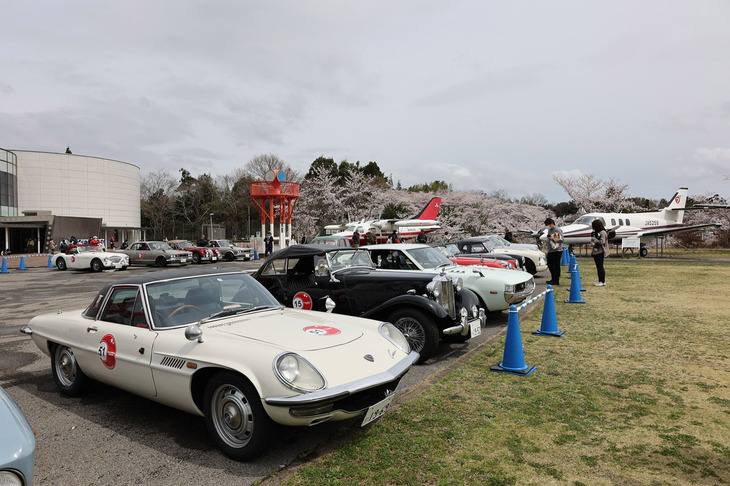 ホンモノの飛行機が並ぶ航空科学博物館の屋外展示スペース近くに車両を止めて、参加者は館内にスタンプをもらいに行く。1969年「マツダ・コスモスポーツ」、1952年「MG TD」、1975年「トヨタ・セリカ」など。