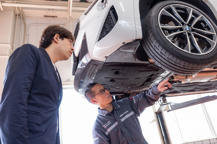 車両をリフトアップし、タイヤに関する点検の手順などを説明するMotoren Saitamaさいたま東のサービス部メカニック永留成浩氏（右）。普段見ることのない車両の下まわりに、サッシャさんも興味津々。