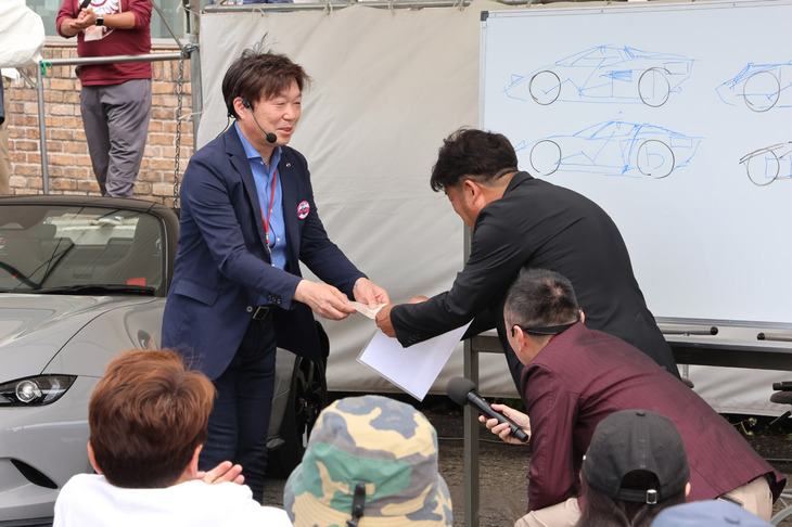 2025年で60歳を迎えた中山 雅さん（写真向かって左）は、この6月でのマツダの退社を公表。軽井沢ミーティング実行委員会の委員長、高橋優一さん（同右）から、愛のある表彰状を授与された。マツダからは離れるが、今後も「軽井沢ミーティング」にはいちロードスターオーナーとして参加することを宣言。会場からは大きな拍手が送られた。今後は新しい仕事に取り組みたいとのことだが、予定は未定とのこと。これからの中山さんの活躍にも注目だ。