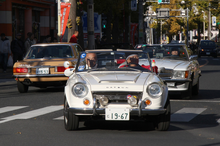 大学通りから谷保駅前のロータリーに入ろうという1967年「ダットサン2000ロードスター」（SRL311）。国内名称「フェアレディ2000」（SR311）の北米仕様である。後方はコードナンバーR107の「メルセデス・ベンツSL」、すれ違っているのも同世代の「同SLC」だ。