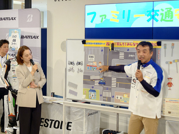 ステージでは、「ファミリー交通安全教室」を開催。これは、道路や駐車場などに潜む日常の危険を親子ともに認知してもらい、安全に過ごすための交通ルールを学ぼうという試みで、講師をレーシングライダーである山口辰也さん（写真右）が務めた。山口さんは、全日本ロードレース選手権ST600シリーズチャンピオン（2010年）などの実績を持つライダーだ。