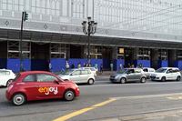 後方のポルタ・ヌオーヴァ駅はただいまファサードを修復中。手前を走り去るのはカーシェアリング「エンジョイ」の「フィアット500」。