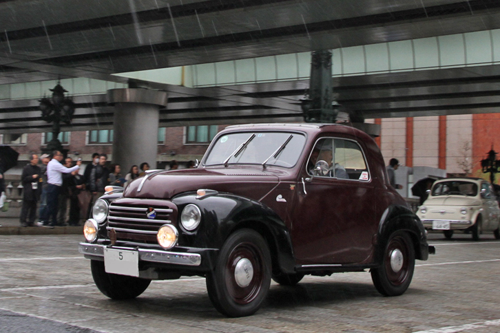 写真に雨粒が写る中を行く、先に後ろ姿を紹介した1953年「フィアット500C」。「5」で始まる陸運支局名がなかった時代の東京ナンバーを付けている。