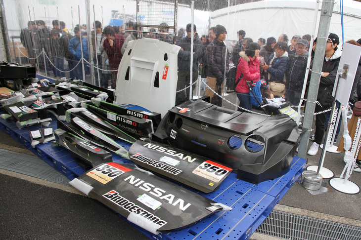 これも毎回好評の、実際にニスモのマシンに使われたパーツ類などを販売するガレージセール。右奥の「GT-R」用CFRP（カーボンファイバー）製リアカウル（40万円）やその隣のボンネット（8万円）は、撮影した午前9時前の時点ですでに売約済みだった。混乱を避けるため、こちらも入場制限されており、後ろに見えるのは順番待ちの来場者の列。