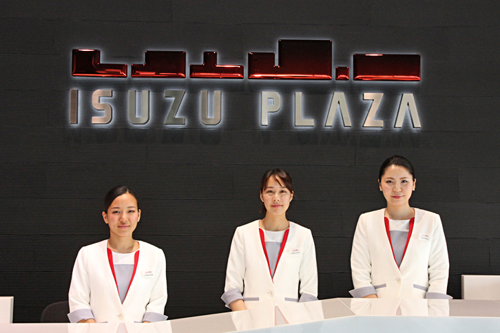 いすゞプラザの受付にて、案内スタッフ。壁面に貼られたロゴマークの「ISUZU PLAZA」の上にあるグラフィックは、建物が並ぶ街の遠景と、いすゞが作るトラックやバスなどのフォルムの双方をモチーフに抽象化。社会といすゞのつながりを象徴しているというが、意図が明快で、かつ的確に表現された秀作だと思う。