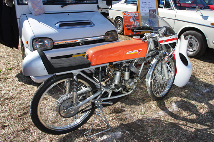 後ろにある「ダイハツ・ミラ ウォークスルーバン」に積まれてきた1963年「トーハツ105Y」。トーハツ（東京発動機から命名）はかつて二輪で名をはせたメーカーで、1964年に倒産。会社再生後は今日までマリーンエンジンや消防ポンプのメーカーとして存続している。二輪メーカーとしてはモペッドの「ランペット」などで知られるが、これは空冷2ストローク2気筒50ccエンジンを積んだ貴重なワークスレーサー。