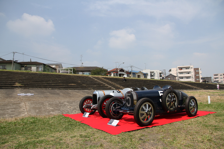 丸子橋付近の多摩川河川敷に残る多摩川スピードウエイのグランドスタンド跡を見上げる場所に展示された、1926年「ブガッティT35C」（手前）と24年「カーチス号」。（除幕前の）記念プレートは、画像左側中ほどに見えるチェッカードフラッグの下にある。