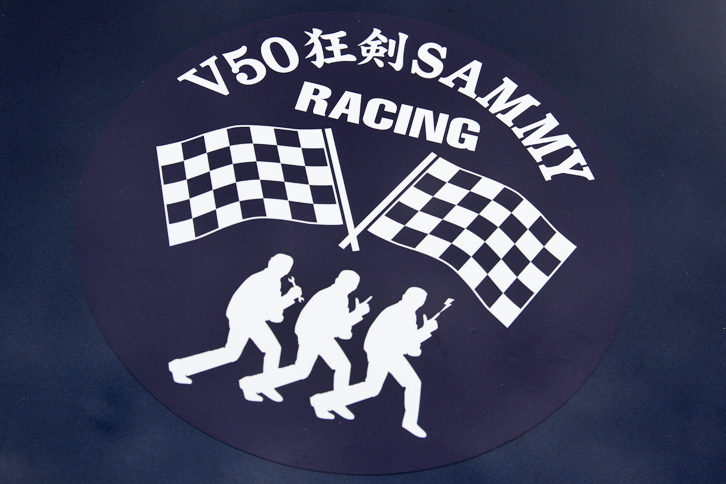トランクリッドに貼られたV50狂剣SAMMY RACINGのステッカー。ボディーサイドにも描かれた3人のシャドウは、中央がおなじみのポーズをキメた剣さん、左側がスパナを持つ内田さん、そして右側は指先から稲妻を放つ桑島さんと、これまた芸が細かい。