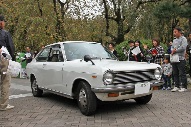 ギャラリーが笑顔で見送るのは、前出の花井 覚さんの初代「ダットサン・サニークーペ」。本人は助手席で、贈られた特別賞の盾を掲げている。