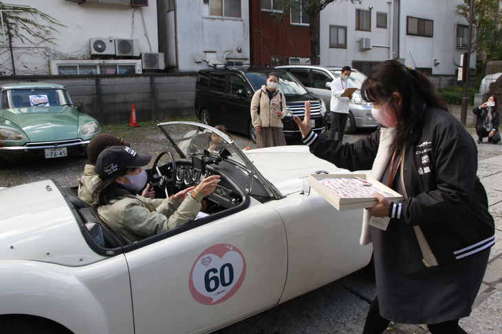スタッフからおみくじを受け取ったのは、1960年「MGA」のコドライバー。後方に見える「シトロエンDS」はスタッフの移動用。