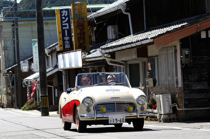これも大野宿を行く、輸出専用車の1960年「ダットサン・フェアレディSPL212」。前日にロケハンとしてコースを一周した際に、個人的に最も印象に残ったのが後方に見える“カンコー学生服”の看板だった。