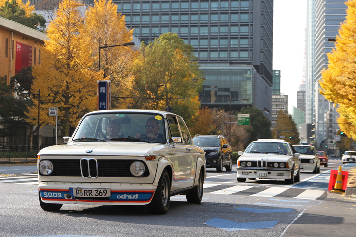 都道301号白山祝田田町線に面した日比谷公園 霞門の交差点を通過する「BMW 2002ターボ」。同じBMWの「M635CSi（M6）」、さらにもう一台の2002ターボが続く。参加車両は横断歩道手前の停止線でトリップメーターをゼロに戻すよう指示された。