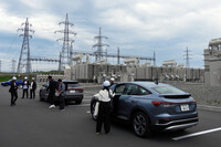 変電所に到着し、クルマを降りる報道関係者。奥に見えるのは巨大な変圧器で、送電線を通る6万6000Vの電気は2200V、610Vと段階的に降圧してバッテリーに蓄電。放電の際には逆の過程を経て送電線に送られる。