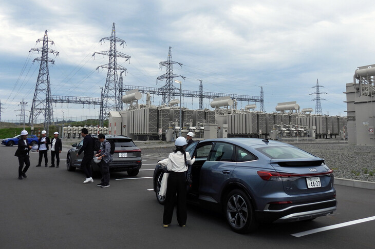 変電所に到着し、クルマを降りる報道関係者。奥に見えるのは巨大な変圧器で、送電線を通る6万6000Vの電気は2200V、610Vと段階的に降圧してバッテリーに蓄電。放電の際には逆の過程を経て送電線に送られる。