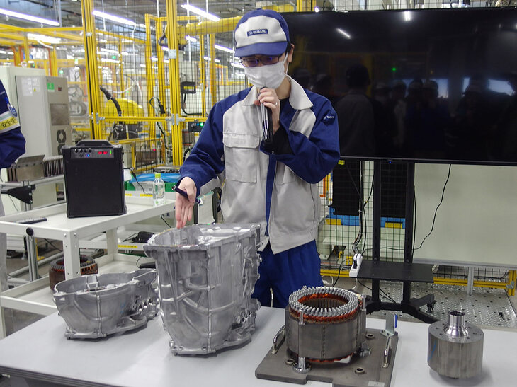 駆動用モーターとそれを内包するケースの組み立てについて説明する工場スタッフ。ケース（写真向かって左から2番目）の内部に、駆動用モーターのステーター（同3番目）とローター（同右端）を組み込むかたちとなる。