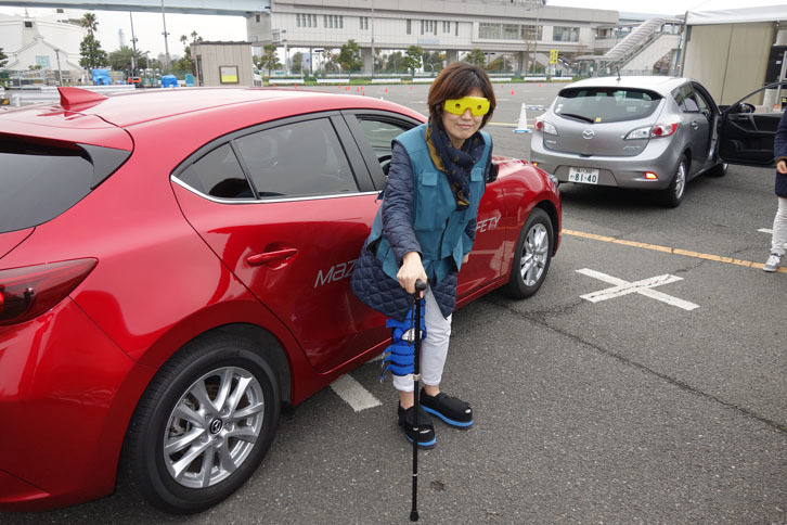 高齢者体験中の筆者。視野を左右100度に制限するめがね、3kgの重りの入ったベスト、ひざのねじれる可動域を制限するサポーター、思ったように足が上がらないことを再現する厚底靴を装着している。