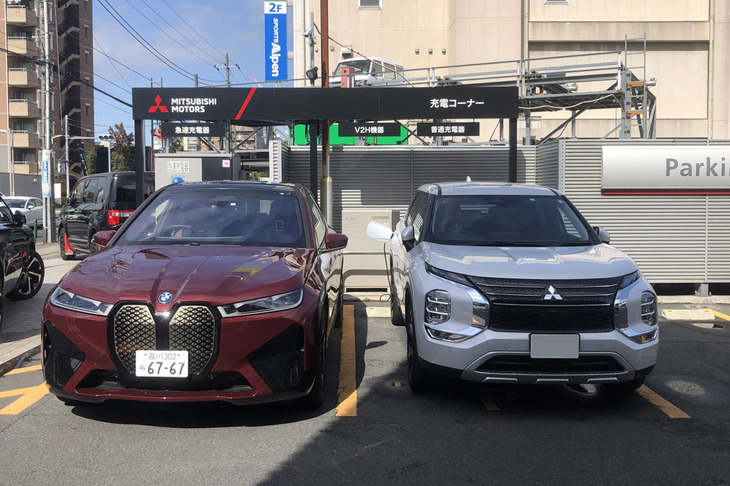 近所の三菱ディーラーで「iX」の充電をすることに。三菱車ではないので開店前にこっそり済ませる作戦だったが、急速充電スペースにはディーラーのPHEV「アウトランダー」が止まっていた。写真は移動してもらったアウトランダー（右）とiX（左）の2ショット。やはりiXの大きさが際立つ。