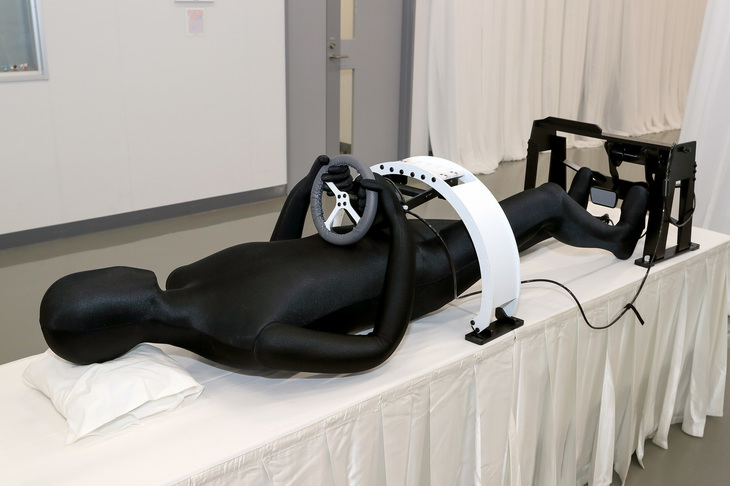 こちらの展示はなにかというと、運転操作中の脳の活動を解析する際に用いる機器である。被験者はこの機器を使い、fMRIの撮影装置の中で寝っ転がりながらドライビングシミュレーターを操作する。