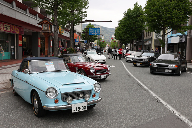 左手前から1964年「ダットサン・フェアレディ1500」と1966年「フェアレディ1600」、右手前に1992年「日産スカイラインGT-R」などが並んだコーナー。中央奥にごく小さく善光寺が見える。