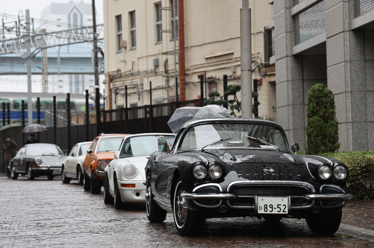 カーナンバー100番以降の汐留西公園に入りきらない参加車両は、いったん近隣の有料駐車場まで連なって移動。1962年「シボレー・コルベット（C1）」を先頭とするその列に……。