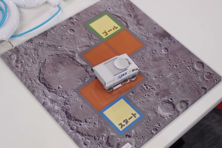 写真中央のミニチュア月面探索車を、自らのプログラミングをもとに操作し、月面ボード（台紙）に記された予定のコースを完走できればミッションクリア。