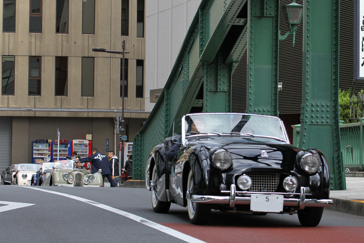 柳橋を中央区東日本橋方面に渡る、「5」で始まる陸運支局名がなかった時代の東京ナンバーを付けた1955年「トライアンフTR2」。後方のスタンプポイントには「ジャガーXK120」や「トライアンフTR3」などが並んでいる。