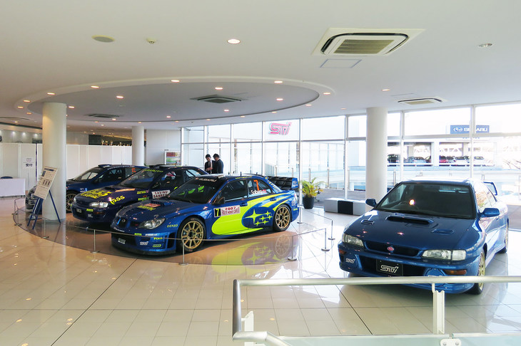 見学会で報道陣を真っ先に迎えてくれたのは、コンプリートカー「インプレッサ 22B-STiバージョン」と歴代のラリーカー。……ただ、このエリアはスバルのショールーム側で、「STIギャラリー」の敷地にはあらず（ギャラリーは写真奥に見える白い仕切りの向こう側）。今だけの特別展示だったようだ。