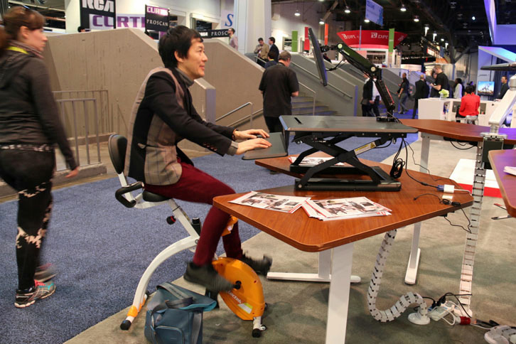 長時間の座り仕事による健康被害を軽減することから、アメリカで近年注目されている昇降デスク。筆者の大矢アキオ（写真）が試しているのはLoctek社のもので、椅子がエアロバイクを兼ねるタイプ。