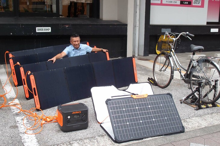 筆者が抱えているのは、最大出力200Wの折り畳み式ソーラーパネル「SolarSaga 200」。人に対しての大きさは写真のとおり。これをキャンプ場でいくつも広げたら……。なにやらスパイ組織の秘密通信基地っぽくて、カッコいいだろう！ 「SolarSaga 200」を4枚組み合わせて使えば、モバイルバッテリー「1000 Pro」を最速1.8時間で充電可能。それらにアダプター2個を加えたセットは「ソーラージェネレーター1000 Pro」として50万5800円で販売される。ちなみに1000 Proの単体価格は14万9800円で、「SolarSaga 80」（写真手前）1枚とのセットは17万8600円。