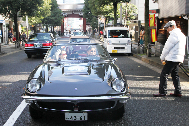 信号待ちしている1965年「マセラティ・ミストラル スパイダー」のドライバーと通りすがりの人が笑顔で言葉を交わす。