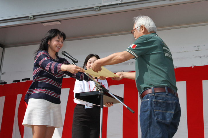 閉会式を前に、参加者による人気投票の上位入賞車をはじめ各種の表彰が行われた。プレゼンターを務めたのは、ゲストであるモータージャーナリストの竹岡 圭さん（写真左）。