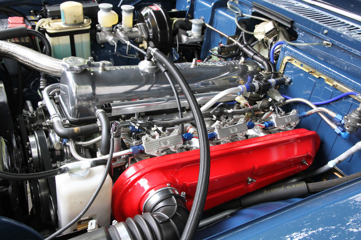 エンジン本体は同じとはいえ、大胆なことをする人もいるもんだ……と思ってよく見たら、たしかにエアクリーナーとインテークマニホールドはS54B用だが、それらの間にあるのはウエーバーではなかった。それどころかキャブでもなく、インジェクションなのだ。オーナーいわく、往年の吸気系チューニングパーツブランドであるSKサンヨー製のスロットルボディーや汎用（はんよう）インジェクターなどを組み合わせたものとのことだが、まるでプリンス純正のような「P」マークを入れるなど、仕上がりが美しい上に芸が細かい。運転席側バルクヘッドには、本来はないバキュームサーボが見えるが、フロントブレーキをオリジナルのドラムからディスクに交換。ラジエーターには電動ファンを装着するなどして、現代の交通環境に対応。そしてこれらの改造は、すべて公認取得済みとのこと。