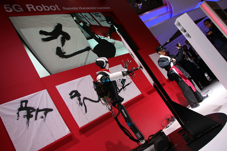 モバイルワールドコングレス2018の会場に展示された、NTTドコモのロボット。右側に立つデモンストレーターの動作を5G通信で受信し、自らの手で書を披露する。