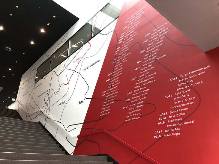 レースカーの開発拠点に入ってすぐの階段には、アウディスポーツの歴史が展示されているほか、逆サイドにはサーキットの場所を示した地図と契約ドライバーの名が連なる。2011年のところにはフレイさんの名も。