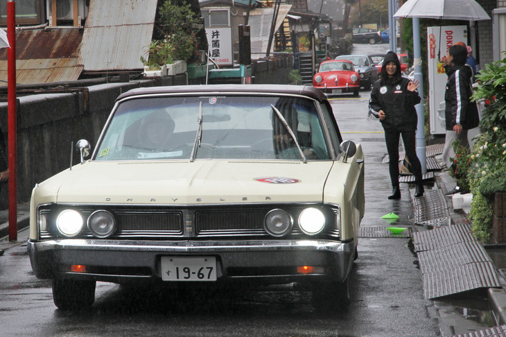 1967年「クライスラー・ニューポート コンバーチブル」。車幅2mに達する、往年のフルサイズの米車が路地を行く。後方には「ポルシェ356」や「BMW 3.0CS」が見える。