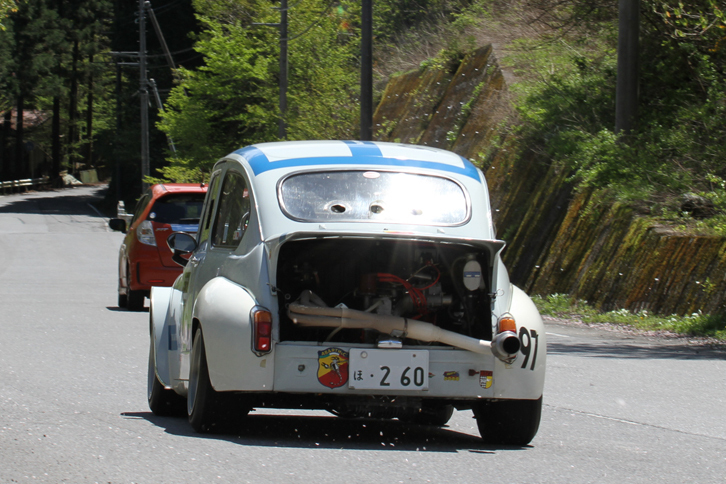 迫力満点の後ろ姿を見せつけながら走り去っていく1964年「フィアット・アバルト1000TCR」。路面に散った、遅咲きの桜の花びらが舞っている。