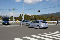 「ITS実験場」に設けられた交差点での、路車間通信の実証実験。実験の主体は手前の「プリウス」で、右折しようとしているが、対向車線の右折待ちの「ハイエース」に遮られた死角から直進車両（クラウン）が出てきて、横断歩道には歩行者（写真右端）もいる。