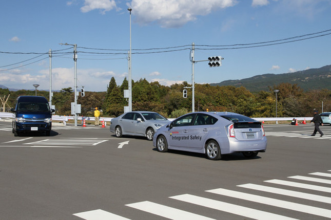 「ITS実験場」に設けられた交差点での、路車間通信の実証実験。実験の主体は手前の「プリウス」で、右折しようとしているが、対向車線の右折待ちの「ハイエース」に遮られた死角から直進車両（クラウン）が出てきて、横断歩道には歩行者（写真右端）もいる。