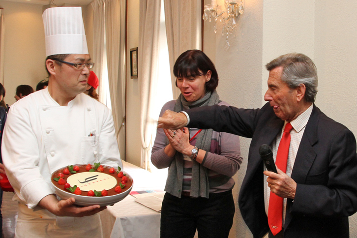 表彰式が行われた施設内のレストランのシェフからフィオラヴァンティ氏に、会社のロゴをかたどったケーキがサプライズとしてプレゼントされた。それを受けてフィオラヴァンティ氏は、早速「指ですくってペロッとなめちゃおうかな～」とおちゃめなジェスチャー。写真中央は、通訳を務めた女性。