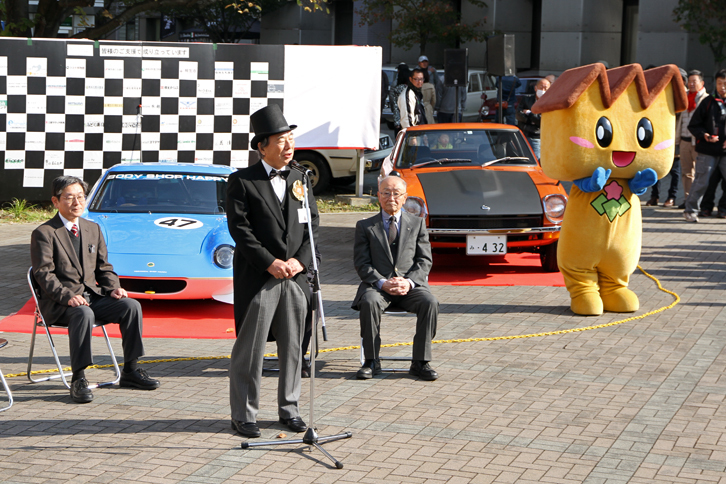 オープニングセレモニー。モーニングコートにシルクハット姿であいさつするのは実行委員長の前原勝良氏。オールド・クラウンを愛好する、地元の内科医院の院長先生である。右端は桐生市の近代化遺産である“のこぎり屋根”の工場をモチーフにしたマスコットキャラクターの“キノピー”。