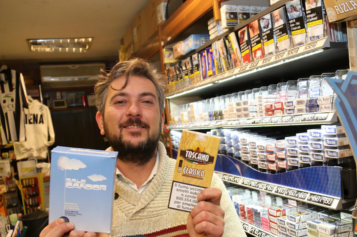 シエナのタバコ店で。店主が手にする葉巻パッケージの警告は、EU基準よりやや小さめ。だが「喫煙は死に至ります」の文字とともに、禁煙相談の電話番号が記されている。後方の箱には患部や、二次喫煙から守るべき幼い子どもの写真が。