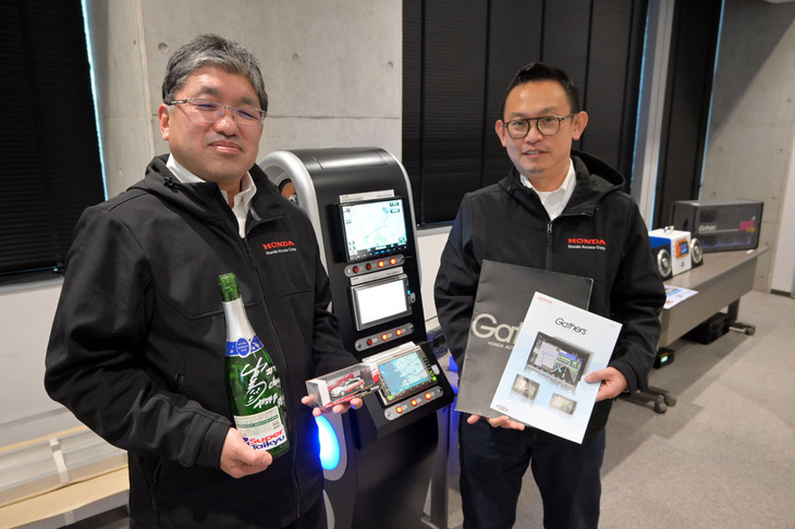 今回、お話を伺ったホンダアクセスの「ギャザズ」商品企画部の大坪浩也氏（左）と加藤智久氏（右）。大坪氏が手にしているのは、スーパーN1耐久レースで、ギャザズがスポンサードしたチームが優勝したときに飲んだシャンパンの瓶。加藤氏が手にするのは、最新と最古のギャザズのカタログだ。