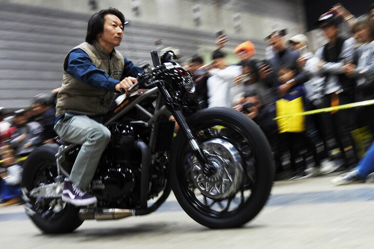 会場のど真ん中の“花道”を通り、著名なカスタムカー／カスタムバイクやゲストが登場する「RIDE IN SHOW」より。自身の手になる「SAMURAI（サムライ）」に乗って登場するカスタムファクトリー「SURESHOT（シュアショット）」の相川拓也氏。