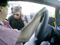 ドライバーズシートの後ろに付けられたウォーキートーキー（トランシーバー）をチェックしに来たインストラクター。先導車から走りながら、またはコース脇に立って指示を出す。ドライバーは、自動車専門誌『Car Graphic』の高平高輝記者。